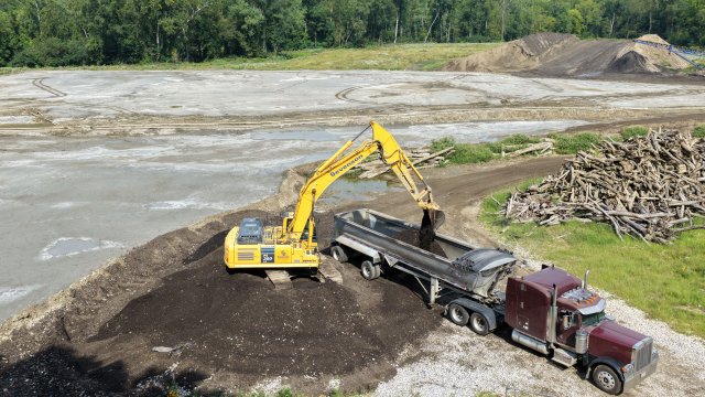 A large yellow excavator scoops brown earthy material into the silver bed of a maroon semi-truck. A gray bare field is in the background along with a large pile of wood debris.