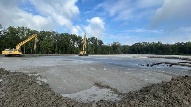 Two large yellow excavators atop a flat bare field with green trees and a blue sky with puffy white clouds in the background. On the right side of the image, two pipes can be seen discharging brown earthen material onto the field.