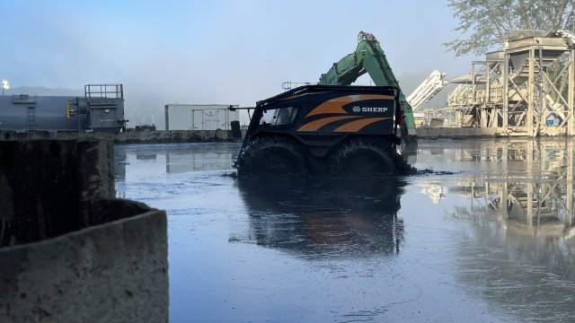 A small black and orange all-terrain vehicle with large treaded wheels drives along the surface of a basin filled with wet, muddy material.