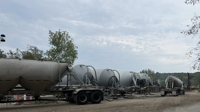 A row of large, gray metal storage tanks with pipes connecting to two delivery trucks. A gray overcast sky fills the background.