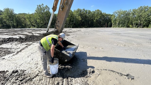  A construction crew member in a gray hard hat and hi-vis green safety vest scoops gray mud from the bucket of a large yellow excavator. The crew member is standing on a flat expanse of gray mud, with green trees and a blue sky in the background.