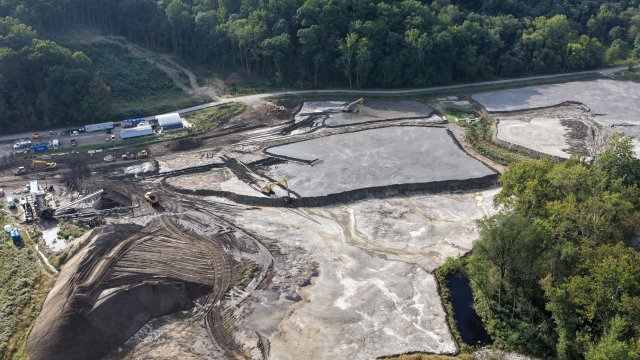 A bird’s-eye view of an open field under construction with mounds of gray earth. Green trees surround the field. 