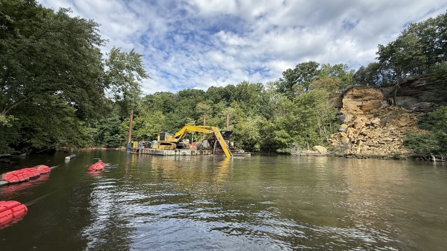 A yellow excavator on a floating barge reaches below the surface of a river. Green trees and a blue sky with puffy clouds fill the background. 