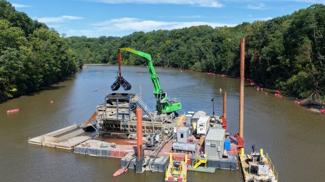 A large green excavator on a barge floating on a river. Green trees and a blue sky fill the background.