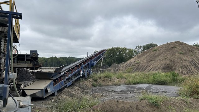 A blue conveyor belt carries brown sandy material up to a large brown mound of sand. A gray overcast sky fills the background. 