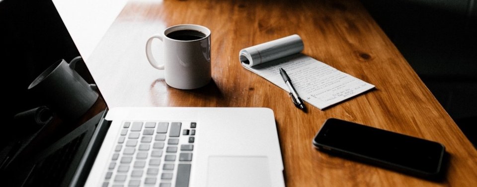 This is a picture of a laptop, a coffee mug, a cell phone, a pen and a pad of paper on a desk