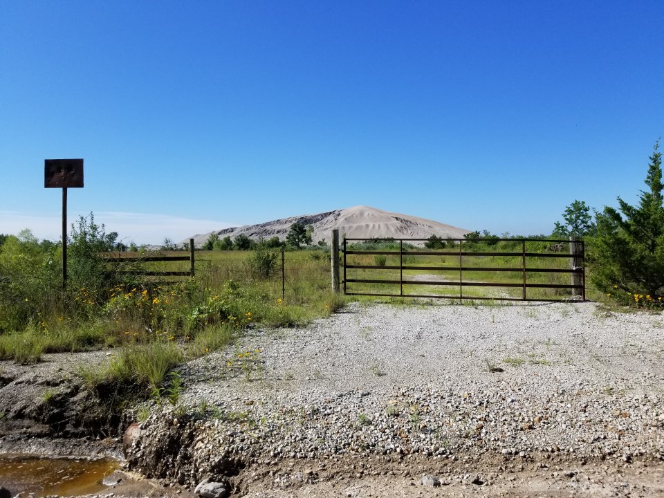 A gravel streambank and gate in front of a vegetated grassy field. A large chat pile sits in the center of the grassy area.