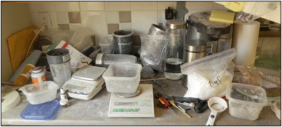 Table surface cluttered with plastic containers, blenders, and plastic bags with a white substance covering the entire scene.