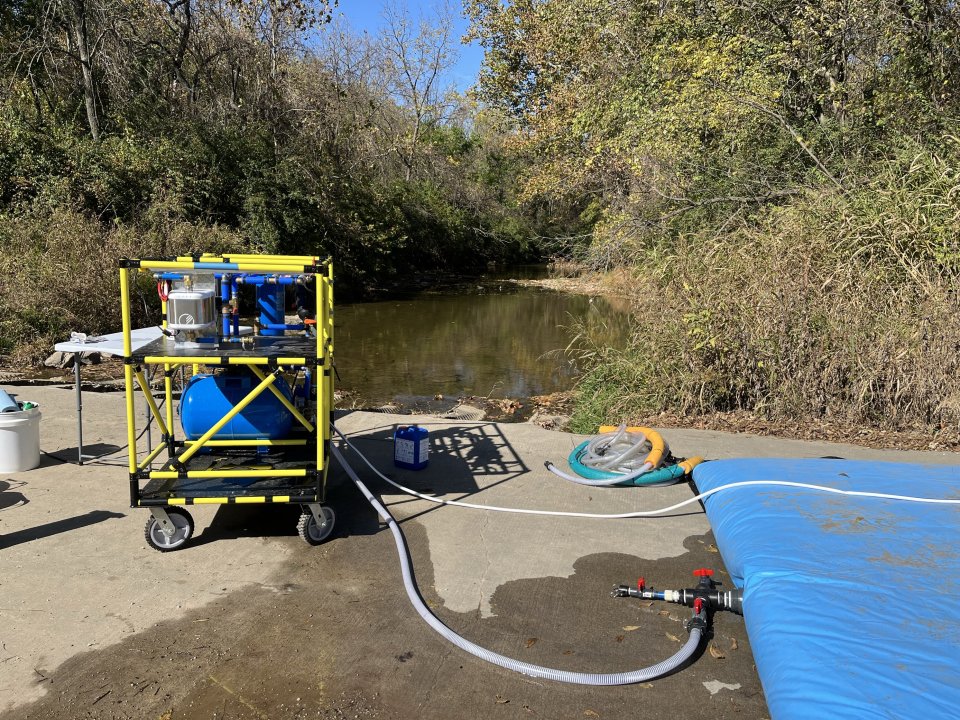 A WOW Cart set up on asphalt with connections leading to the blue bladder tank next to a body of water.