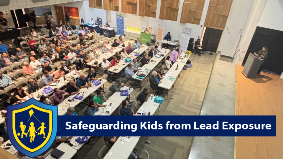 Wide indoor photo of a large group gathered in a conference room with rows of tables, listening to presenters at the front. A banner across the bottom reads “Safeguarding Kids from Lead Exposure.”