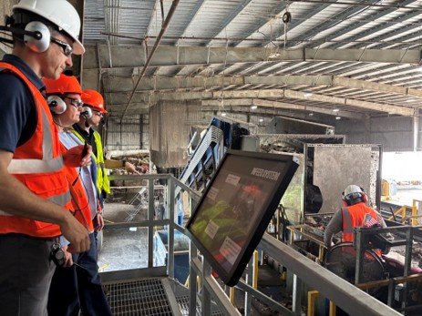 photo of a woman wearing a helmet, safety glasses, hearing protection, next to men in hi viz work vests wearing the same overlooking trash sorting done by machines