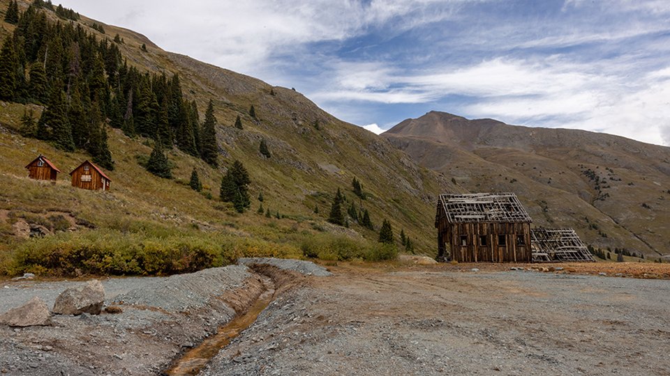 Bonita Peak Mining District area with dilapidated wooden structures on a blue sky cloudy day and pine trees on the mountain ridge.