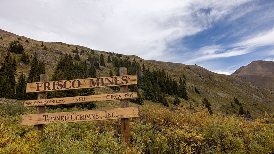 A weathered and worn wooden sign stating Frisco Mines, Circa 1905, Tunnel Comoany Inc in the foreground with cloudy blue sky and moutains with pine trees in the background.