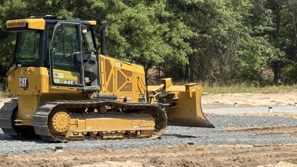 Yellow CAT bulldozer parked on gravel area at a lead service testbed site, with dirt and trees in the background.