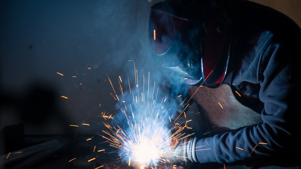 A person wearing a protective mask welds metal; sparks fly.