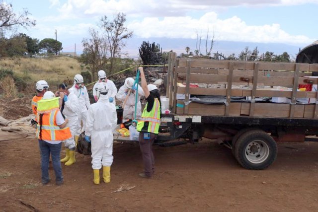 Eight workers, five in Hazmat Suits, three in high visibility vests, prepare for work at the back of a work truck