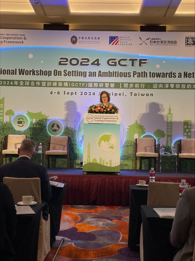 Jane Nishida standing on stage in front of podium giving presentation with 2024 GCTF banner in background.