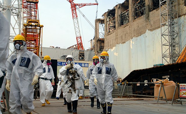 IAEA experts wearing tyvek suits, hard hats, and respirators walking outside of a damaged nuclear power plant station.