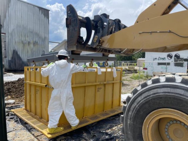 Worker in protective gear oversees the loading of contaminated soil bags into a container in a staging area.