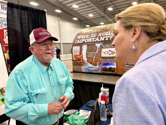 Woman in a jacket with earrings talks to man in hat behind a booth with soil on the table and information about soil health