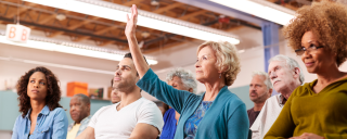 A woman raises her hand while seated among a group of people at a town hall meeting