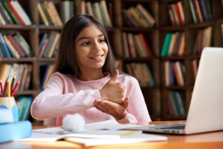 Girl doing sign language while looking at laptop in front of bookshelf.