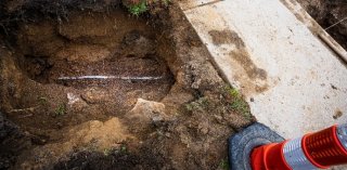 A picture containing a hole in the ground next to a sidewalk and a caution cone. A water pipe is visible in the dirt.