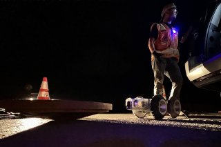 CCTV camera on four wheels next to a manhole cover. There is a cone and a man in the background.