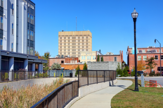 A sidewalk vertically intersects the lower portion of the screen. A building is to the left of it and a grassy area with a black lampost is to the right. The background has medium sized office buildings against a brillian blue sky. 