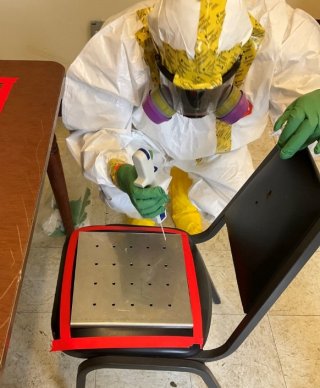 Responder suited up in a Tyvek suit leans over a black chair and deposits malathion onto a coupon on the seat that is marked in a red square.