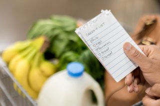 Hand holding a handwritten grocery list over a shopping cart. The cart contains a jug of milk, greens, bananas, and a purse.