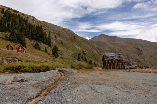 Bonita Peak Mining District area with dilapidated wooden structures on a blue sky cloudy day and pine trees on the mountain ridge.