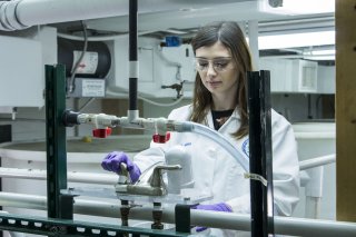 EPA scientist wearing a lab coat and protective gloves and goggles taking water samples from EPA's home plumbing system laboratory.