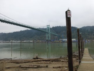 Beach in foreground has large drift wood. Pilings along walkway say No Rishing. A bridge crosses the waterway with hilly landscape on the far side.