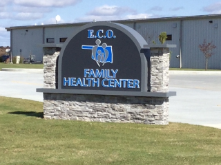A sign for the E.C.O. Family Health Center in front of a parking lot and a large building.