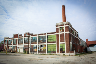 A dilapidated brick industrial building with busted out windows sets in an open parking lot.