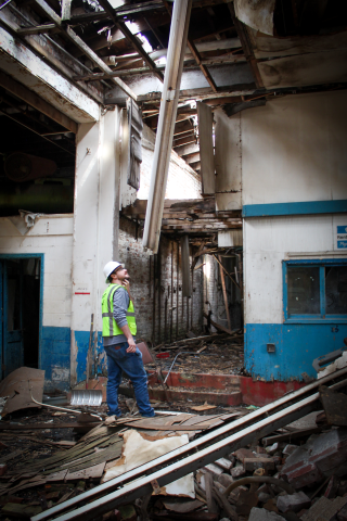 A man, wearing a white hard hat and a reflective safety vest inspects a portion of a caved-in roof in a dilapidated structure.