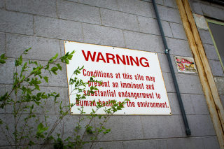 Behind tree growth, a rusting white sign in red letters is nailed into a grey, brick wall and reads: “WARNING – Conditions at this site may present an imminent and substantial endangerment to human health or the environment.”