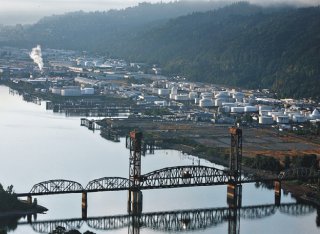 Industrial scene showing steel rail bridge across river with industrial tanks and structures along shore in background.