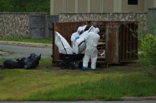 Two people in white tyvek suits putting two white bags of waste into a wheelbarrow while standing in front of an open brown shipping container holding waste.