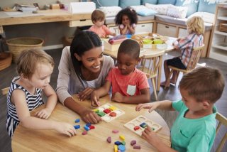 A group of young children work on a puzzle at a small table while a smiling childcare-worker assists.