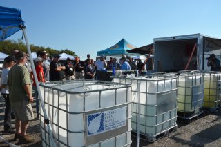 A crowd of people listen to a presentation around four tanks of water.