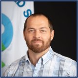 A man with light skin, very short brown hair and beard. He is standing in front of the EPA Flag.