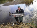 A man wearing a ballcap holds a fish by a small rocky river.