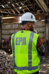 A man, wearing a white hard-hat and a reflective safety vest with the letters “EPA” emboldened on the back, stands in front of a pile of bricks in a dilapidated structure.