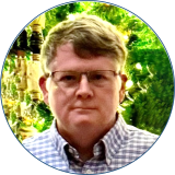 Speaker Joseph Wood is wearing glasses and a checkered button down shirt while standing in front of a green background.
