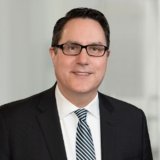 headshot of Dave Ross wearing glasses, black blazer, white, collared shirt, and blue tie.