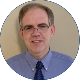 Matthew has dark hair and eyeglasses and is wearing a blue collared shirt with a dark tie