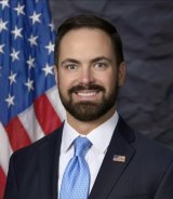 A bearded man in a dark suit and light blue tie. The American flag is in the background.