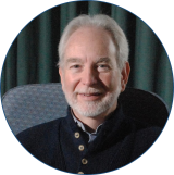 White male with white hair and beard sits at a pale blue chair in front of a dark green curtain wearing a dark blue sweater.